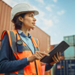 Smiling Portrait of a Beautiful Latin Female Industrial Engineer in White Hard Hat, High-Visibility Vest Working on Tablet Computer. Inspector or Safety Supervisor in Container Terminal.