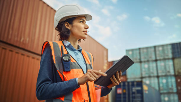 Smiling Portrait of a Beautiful Latin Female Industrial Engineer in White Hard Hat, High-Visibility Vest Working on Tablet Computer. Inspector or Safety Supervisor in Container Terminal. Evolving Gender Roles in Relationships