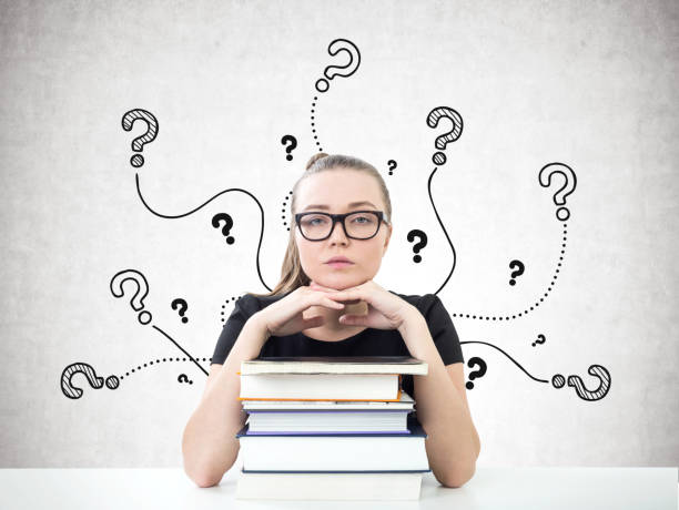 Portrait of a serious young woman in glasses looking forward and sitting with her chin on a stack of books. A concrete wall background with question marks
