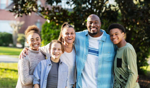 Portrait of a multi-ethnic blended family of five standing together outdoors in their front yard, posing for the camera. The African-American father is in his 40s. Their son and daughter from his prior marriage are standing on the ends. They are 13 year old twins, mixed race African-American and Caucasian. The mother is in her 30s and their youngest son from her prior marriage is 11 years old.