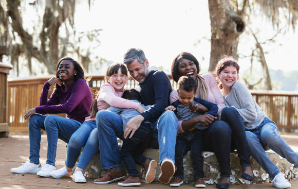 A multi-ethnic blended family playing in the park together on a sunny day. They are sitting together on a deck. The African-American mother and Caucasian father have mixed race twin boys, almost 3 years old. Their daughters and step-daughters are 11 to 15 years old. The mother and girls are smiling and looking, looking at the camera. Blended Family Parenting Tips