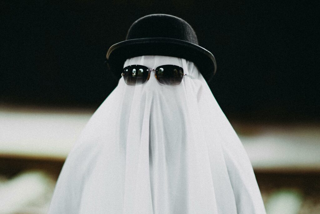 A person with a sheet over their head wearing sunglasses and a black hat.