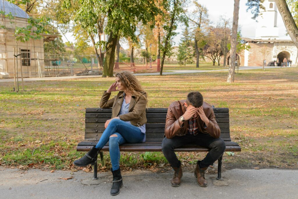 A couple sitting on a park bench after just arguing. The man has his head in his hands and the woman is facing away from him with her hand on her face with her legs crossed.