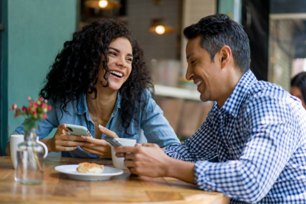 Happy Latin American couple over 40 on a date at a cafe looking at social media on their cell phones and laughing â lifestyle concepts