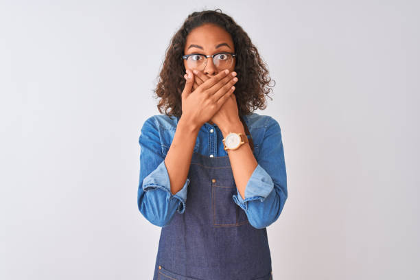 Young woman wearing apron and glasses over isolated white background shocked covering mouth with hands for mistake. Secret concept. Most Embarrassing Thing You’ve Ever Done on a Date?