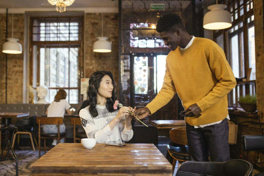 A black man giving a flower to an oriental woman sitting at a restaurant table on their first date.