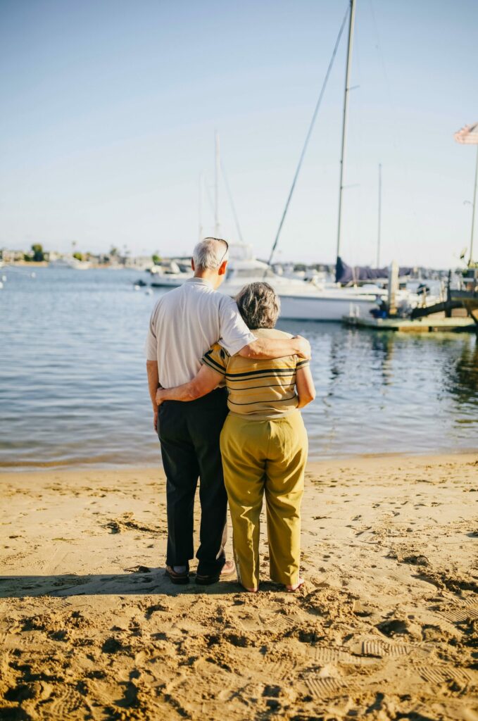 An older couple embracing facing the water and a boat. They are standing on a beach.