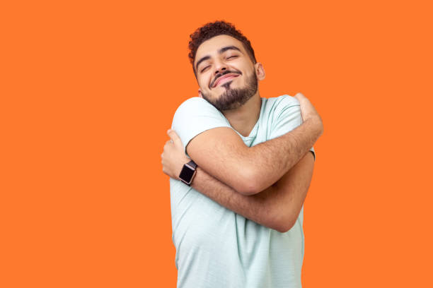 I love myself! Portrait of egoistic brunette man with beard in white t-shirt standing with closed eyes, embracing himself and smiling form pleasure and proud. indoor, isolated on orange background