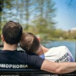 the picture is from the back of two men in a LGBTQ relationship sitting on a park bench looking at a pond. One man has his head on the other mans shoulder. They are both wearing t-shirts that say "No