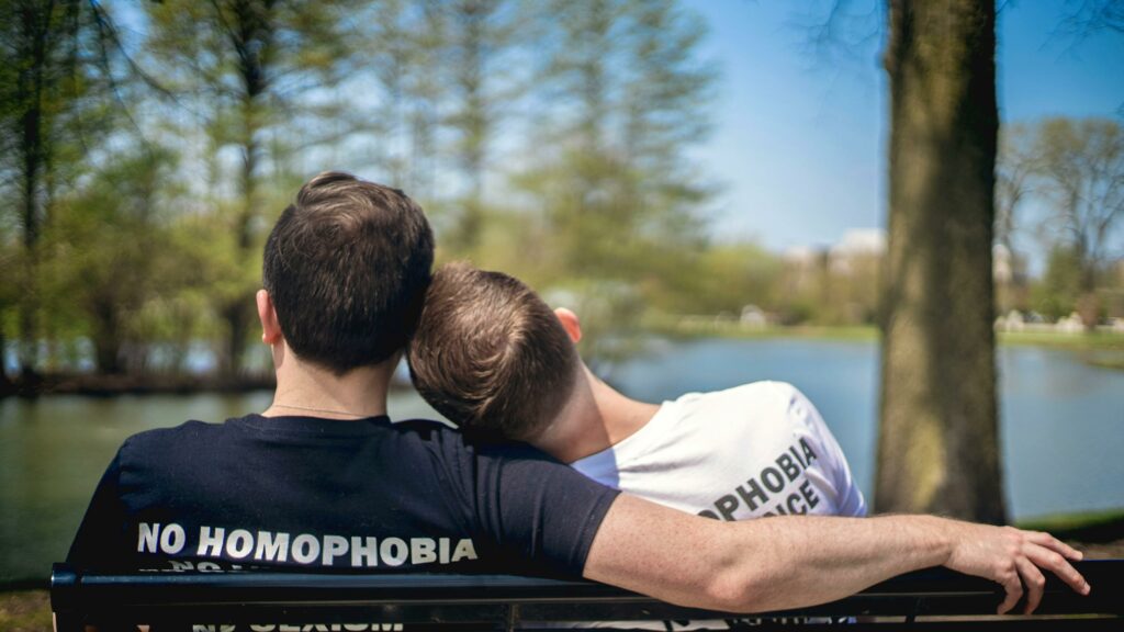 the picture is from the back of two men in a LGBTQ relationship sitting on a park bench looking at a pond. One man has his head on the other mans shoulder. They are both wearing t-shirts that say "No