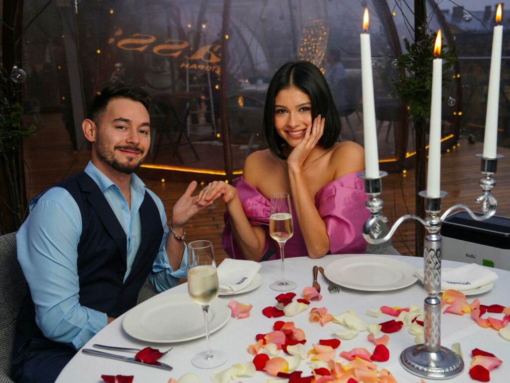 A couple on their first date. They are facing the camera holding hands sitting at a restaurant table. Table has rose petals and candle holder holding three candles lit.