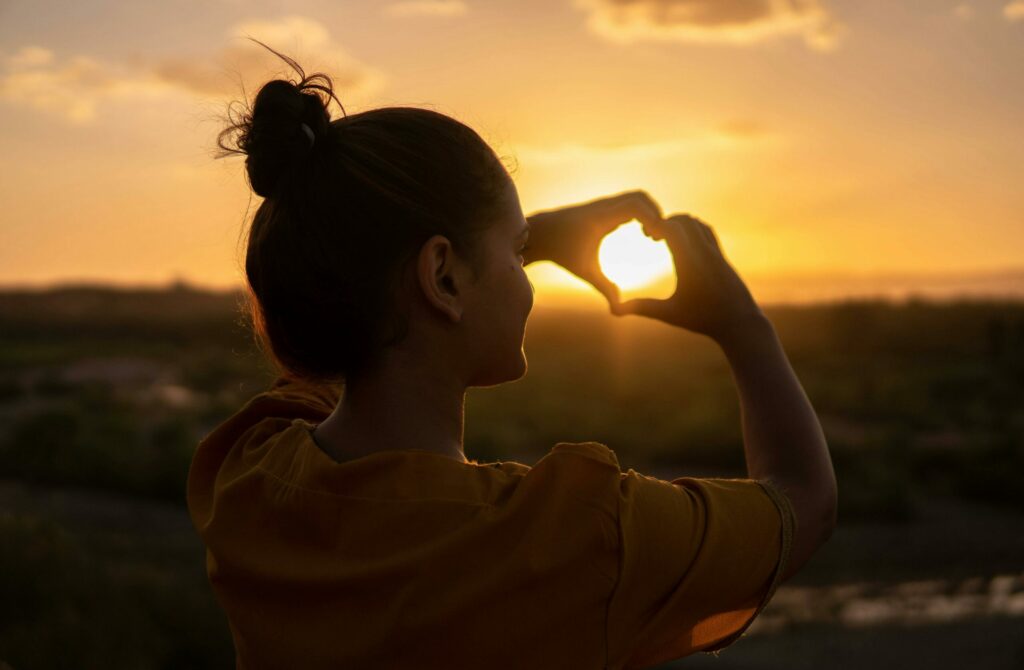 A woman making a heart with her hands facing a sunset and the sun is in the middle of the heart.