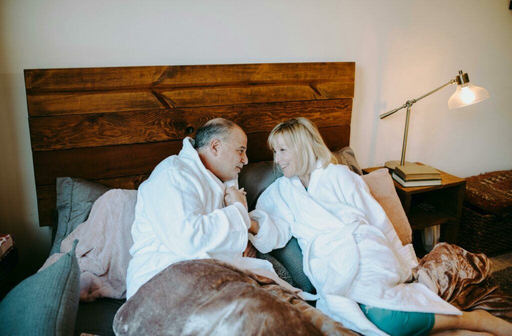 a couple laying in bed with robes on. The man is holding the women hand and their discussing Ways to Improve Sexual Communication